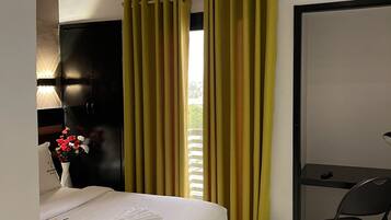 A hotel room with a bed, a desk, a chair, a trash bin, and a window with curtains.
