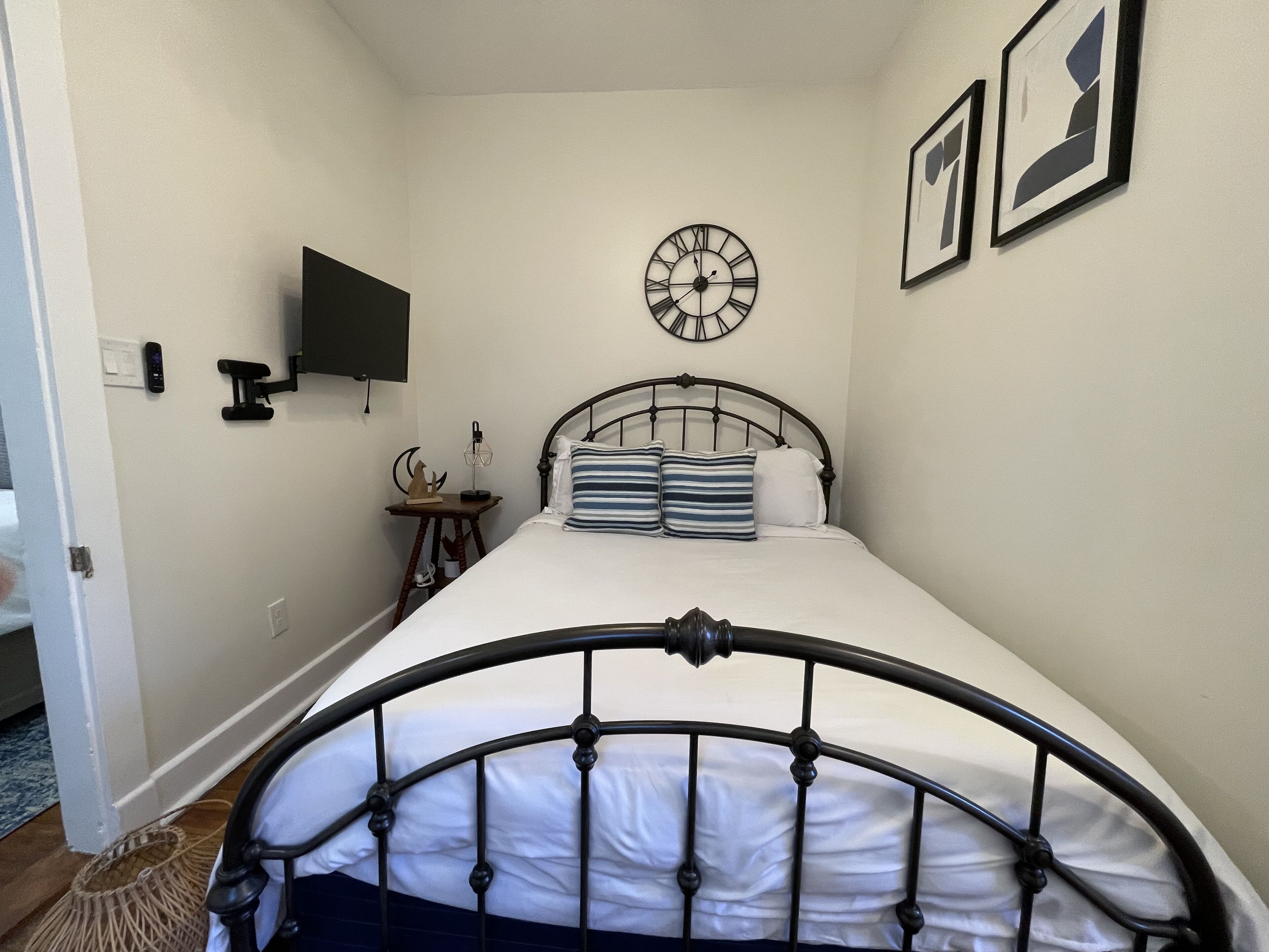 A bedroom with a bed, a wall clock, a bedside table with a lamp, and a wall-mounted TV.