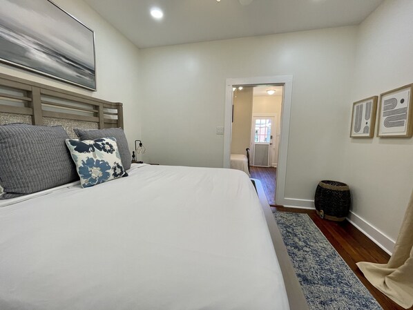 Un dormitorio con una cama grande, una mesita de noche, una lámpara, una puerta que da a otra habitación y una pequeña alfombra.