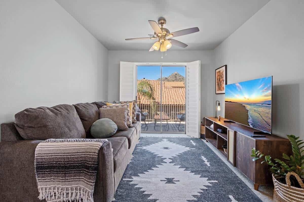 A living room with a sofa, a ceiling fan, a TV, and a view of the outdoors.