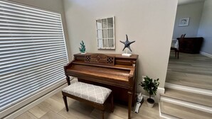 Un piano avec un banc dans une pièce dont le sol est recouvert de carreaux, une fenêtre avec des stores, une petite plante et un miroir accroché au mur.