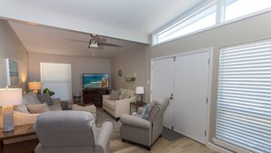 Un salon spacieux doté d’un ventilateur de plafond, d’un téléviseur à écran plat et d’une grande fenêtre avec des stores.