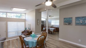 Une salle à manger spacieuse avec un grand ventilateur de plafond, un plancher en bois et une vue sur la cuisine adjacente.