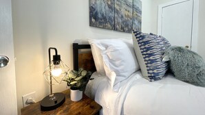Un dormitorio ordenado con cama, almohadas, una lámpara de noche y un cuadro colgado en la pared.