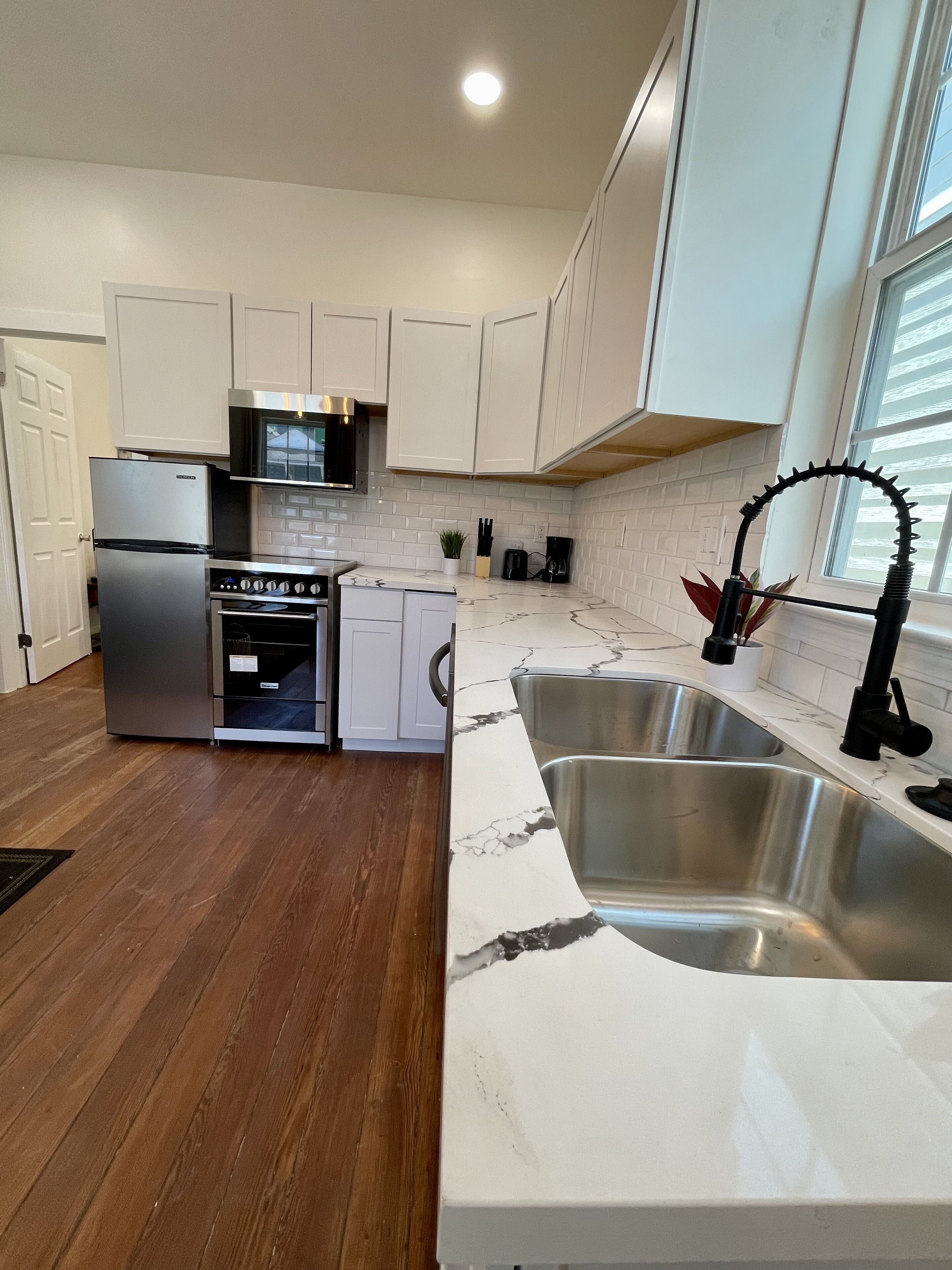 A modern kitchen with white cabinets, stainless steel appliances, a marble countertop, and a large window.