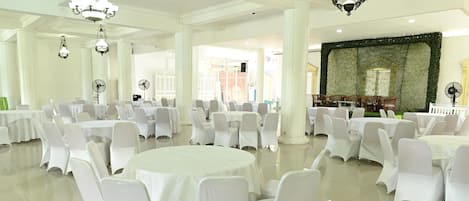 Un salón de banquetes con mesas redondas y sillas blancas, ventanas amplias y candelabros.