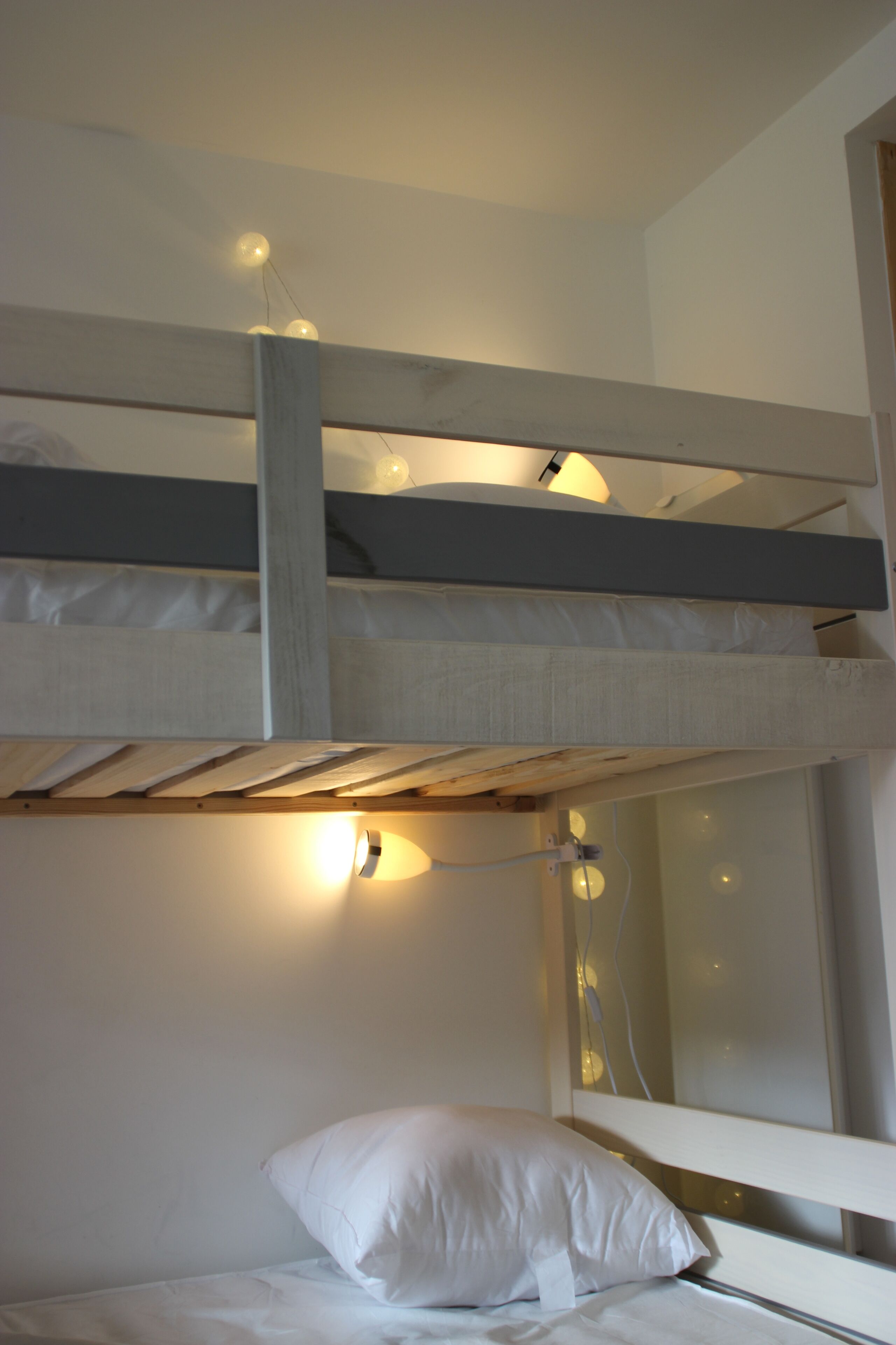 A bunk bed with a white mattress and pillow, a wooden headboard, and a wall-mounted light fixture.