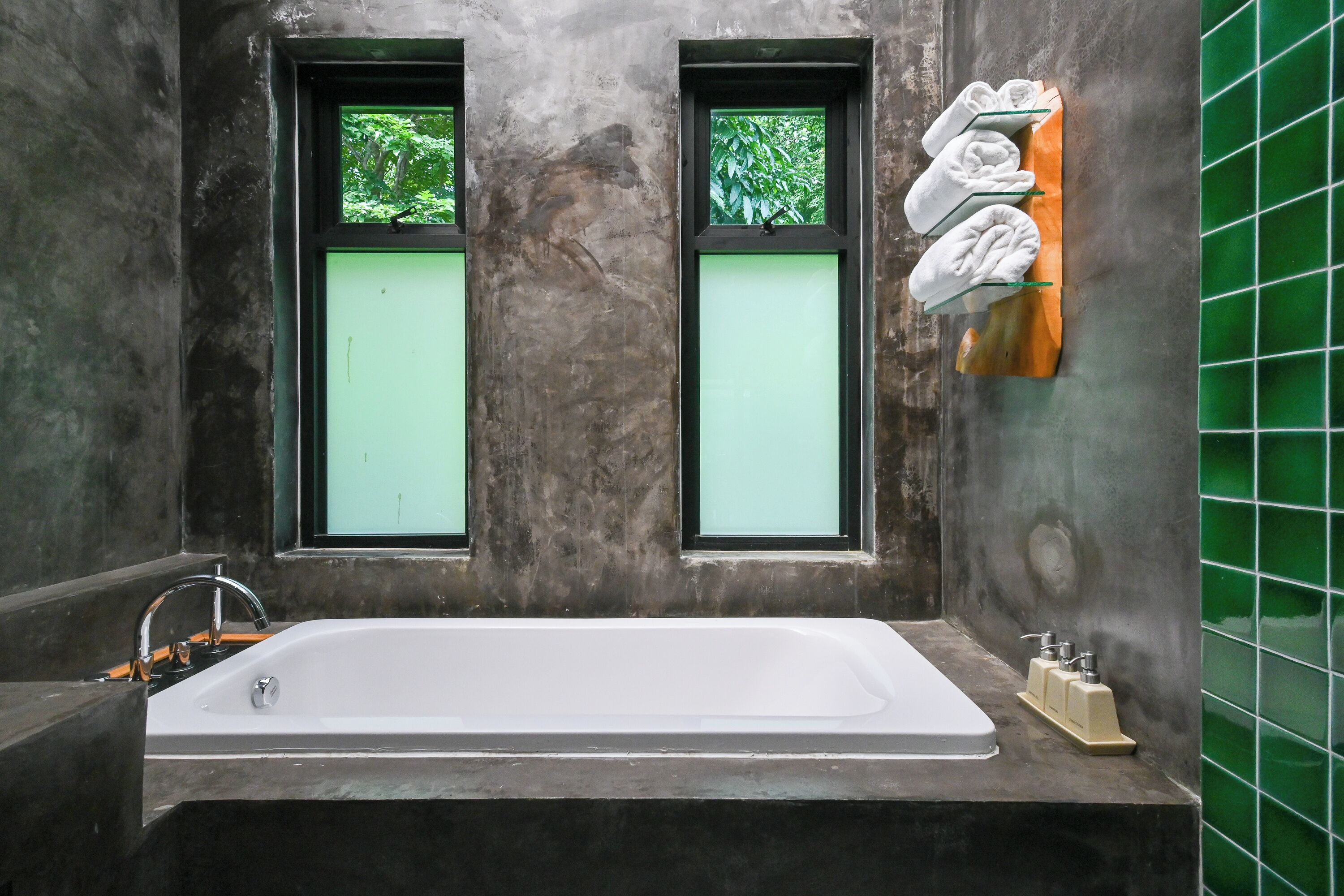 A bathroom with a white bathtub, two windows with green-tinted glass, and a towel rack with folded towels.