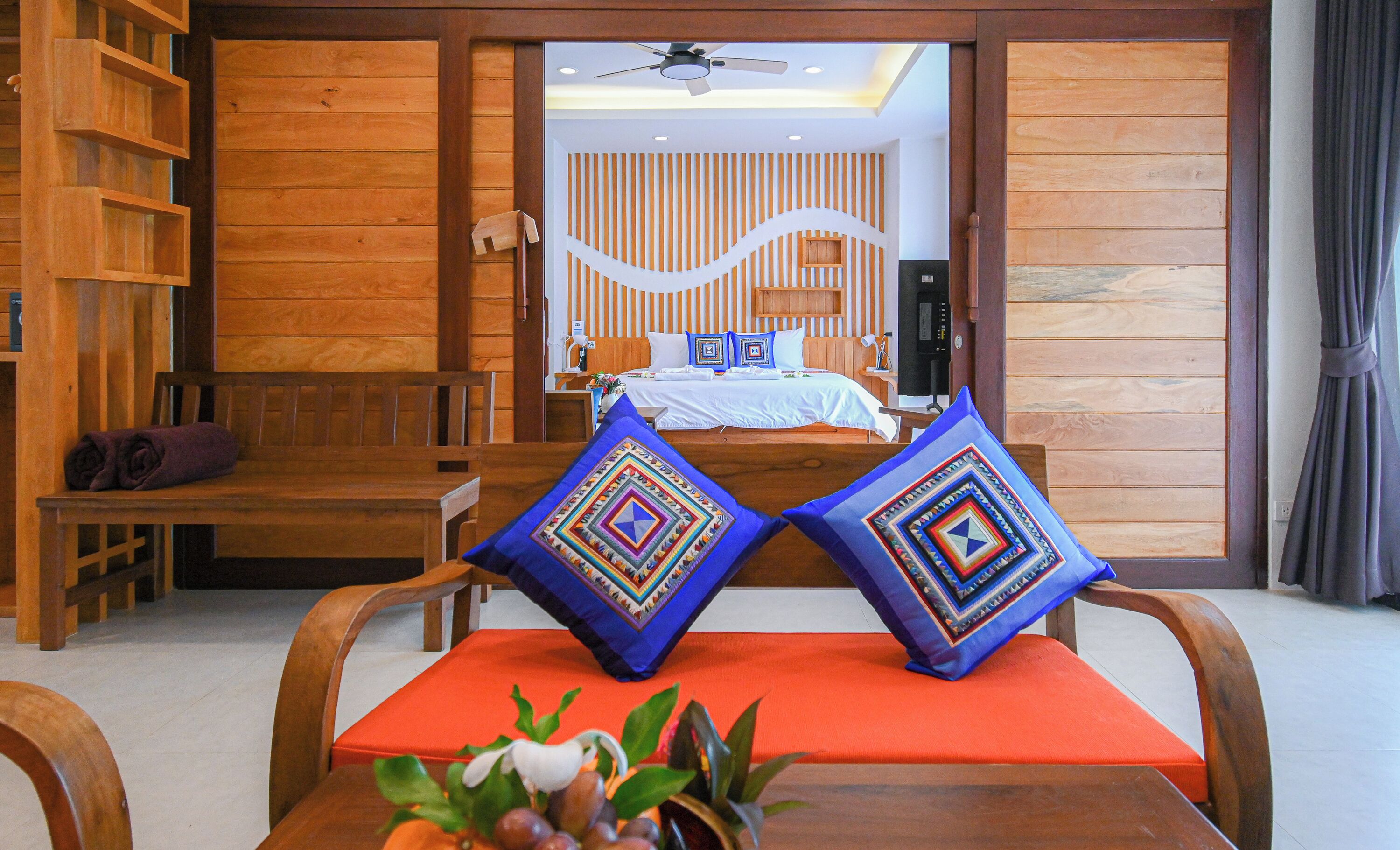 Habitación de hotel con detalles de madera, una cama con ropa blanca, dos almohadones decorativos, un banco de madera y una mesita con una planta.