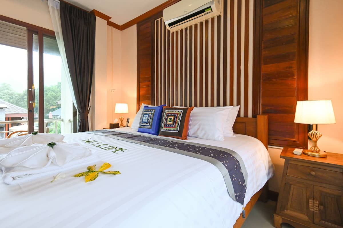 A hotel room with a neatly made bed, a wooden headboard, an air conditioning unit, and a view of the outdoors through a sliding door.