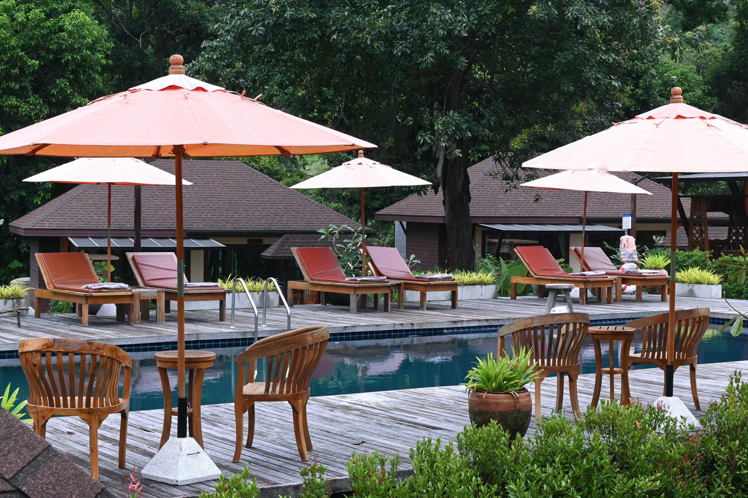 A poolside area with wooden lounge chairs and orange umbrellas, surrounded by greenery and a building in the background.