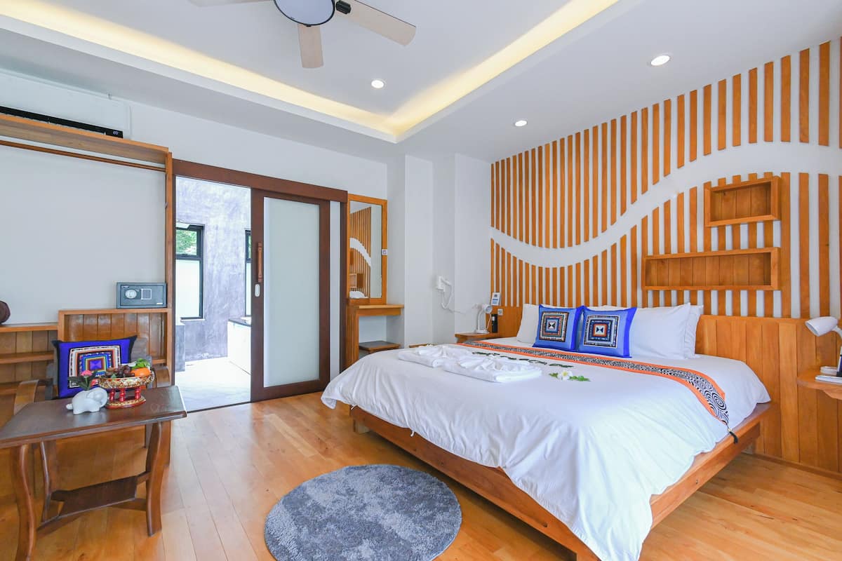 A bedroom with a wooden bed, a desk, and a chair. There is a ceiling fan, a window, and a door leading outside.
