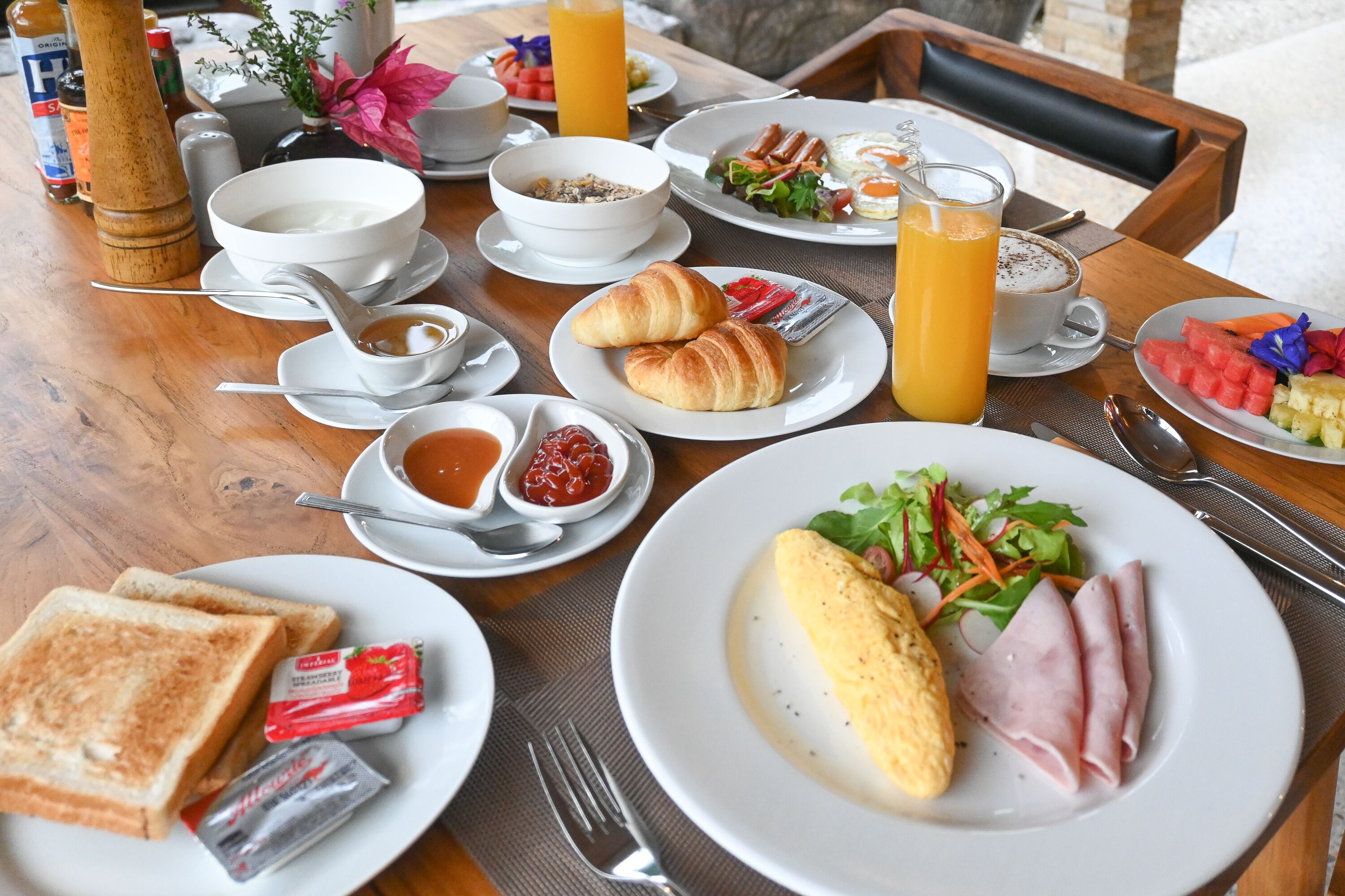 Una mesa de desayuno con cruasanes, tostadas, huevos, jamón y una ensalada de frutas.