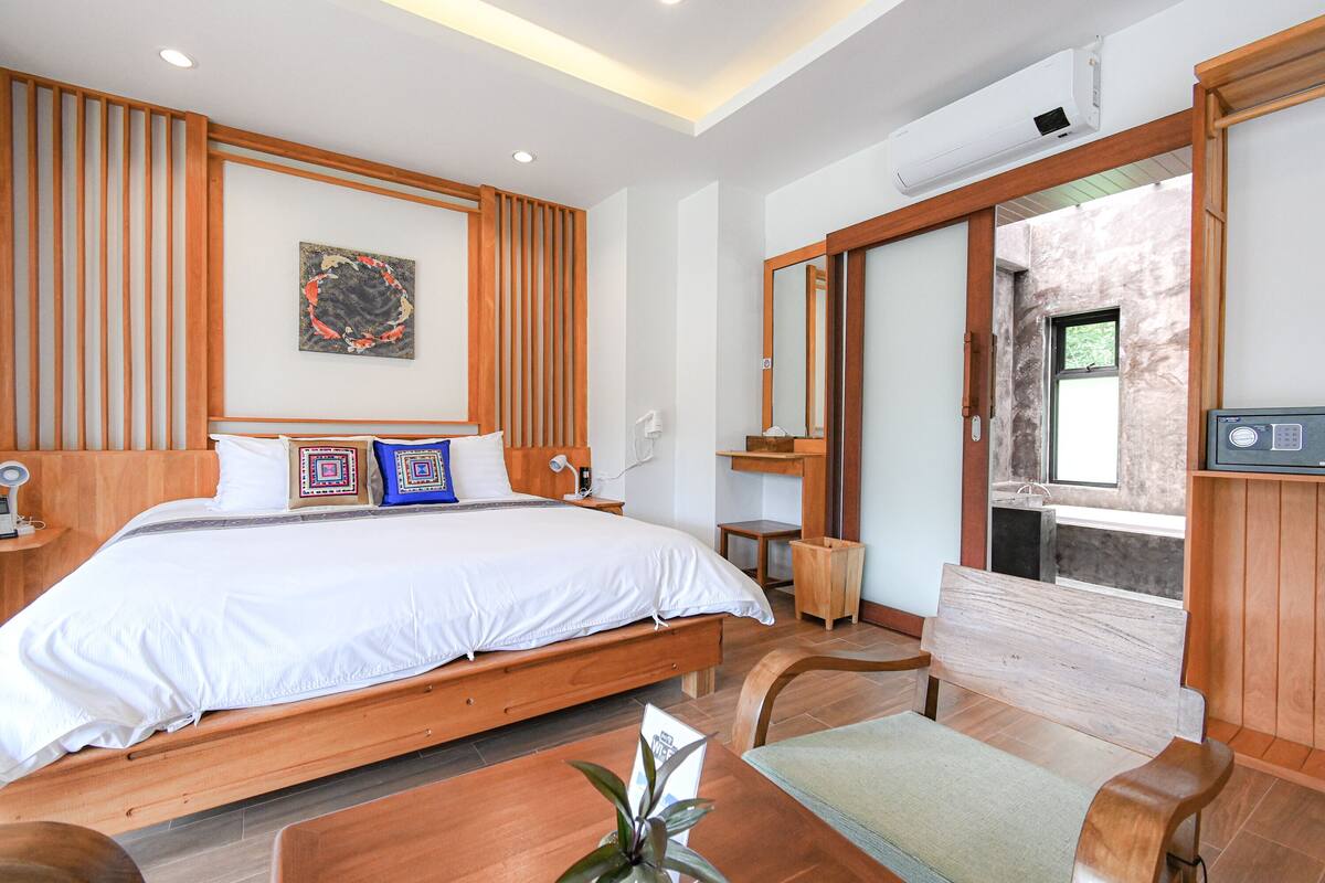 A modern hotel room with a large bed, wooden headboard, a small table, a bench, and a view of the outside.