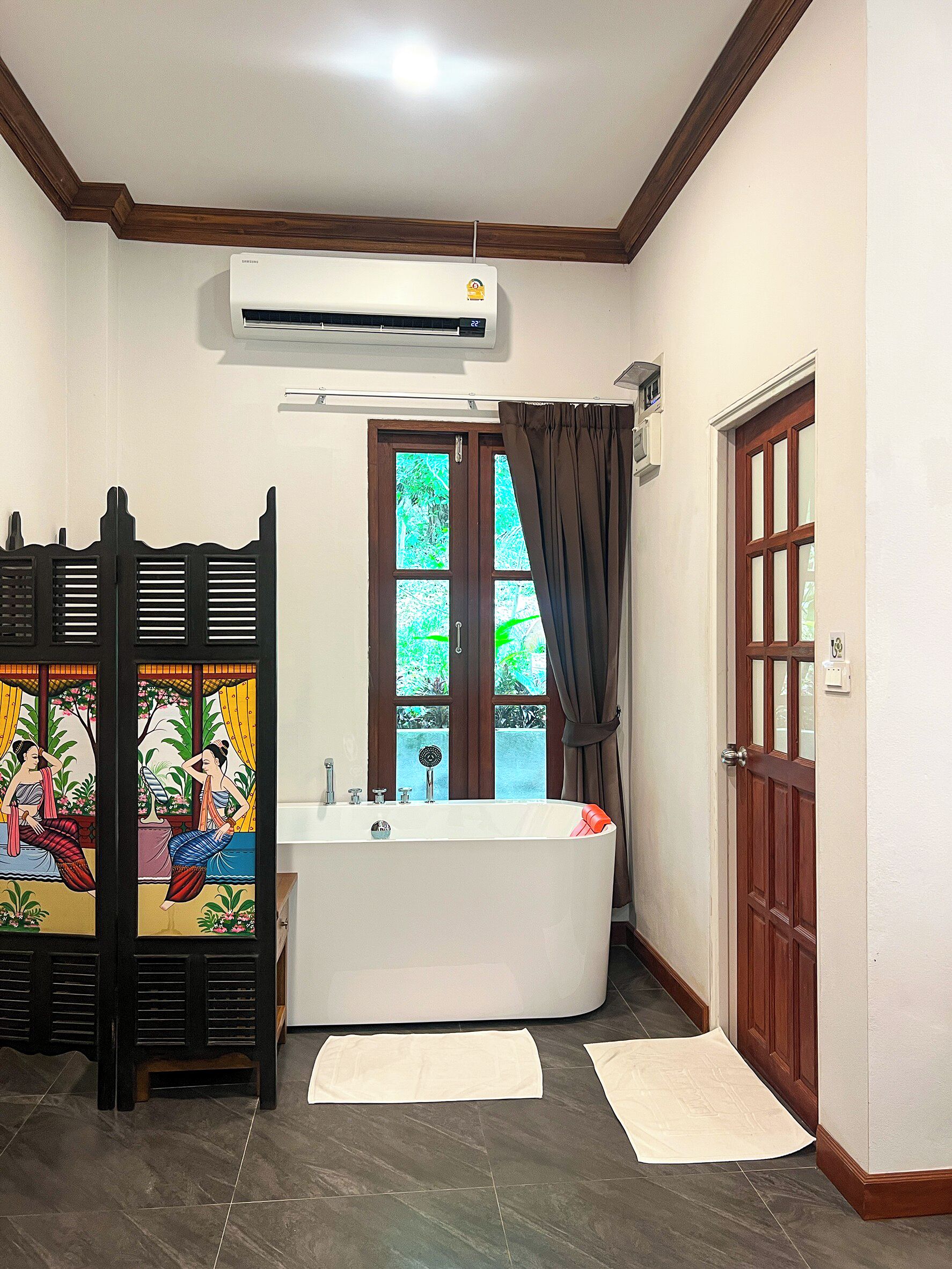 Ein Badezimmer mit einer weißen Badewanne, einem Fenster mit Vorhängen, einer Tür und einer Klimaanlage.