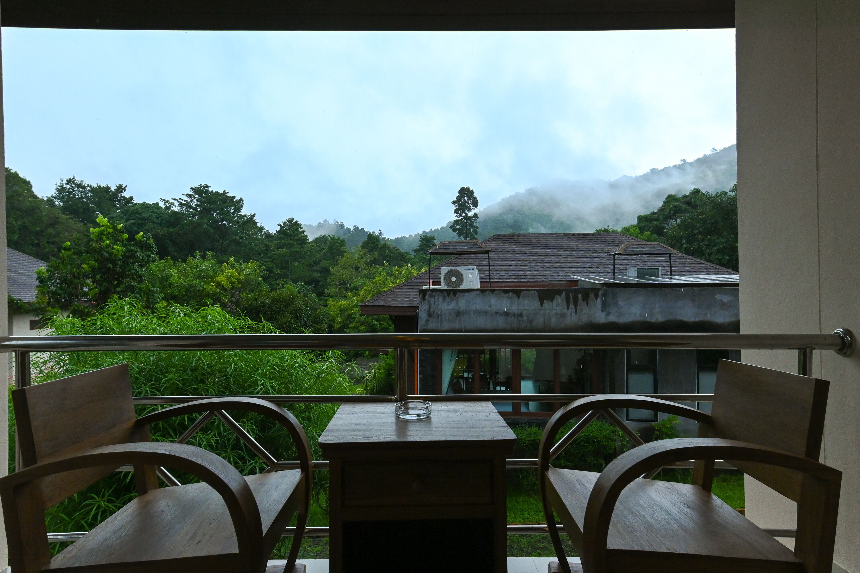 A balcony with two wooden chairs and a small table, overlooking a house and lush greenery with a mountain in the background.
