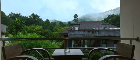 A balcony with two wooden chairs and a small table, overlooking a house and lush greenery with a mountain in the background.