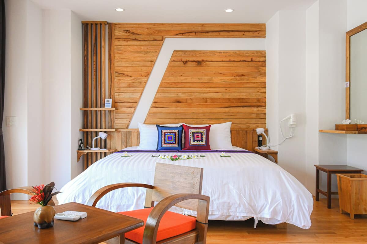 A bedroom with a wooden slat wall, a bed with white bedding, two pillows, a bedside table with a lamp, and a chair with an orange cushion.
