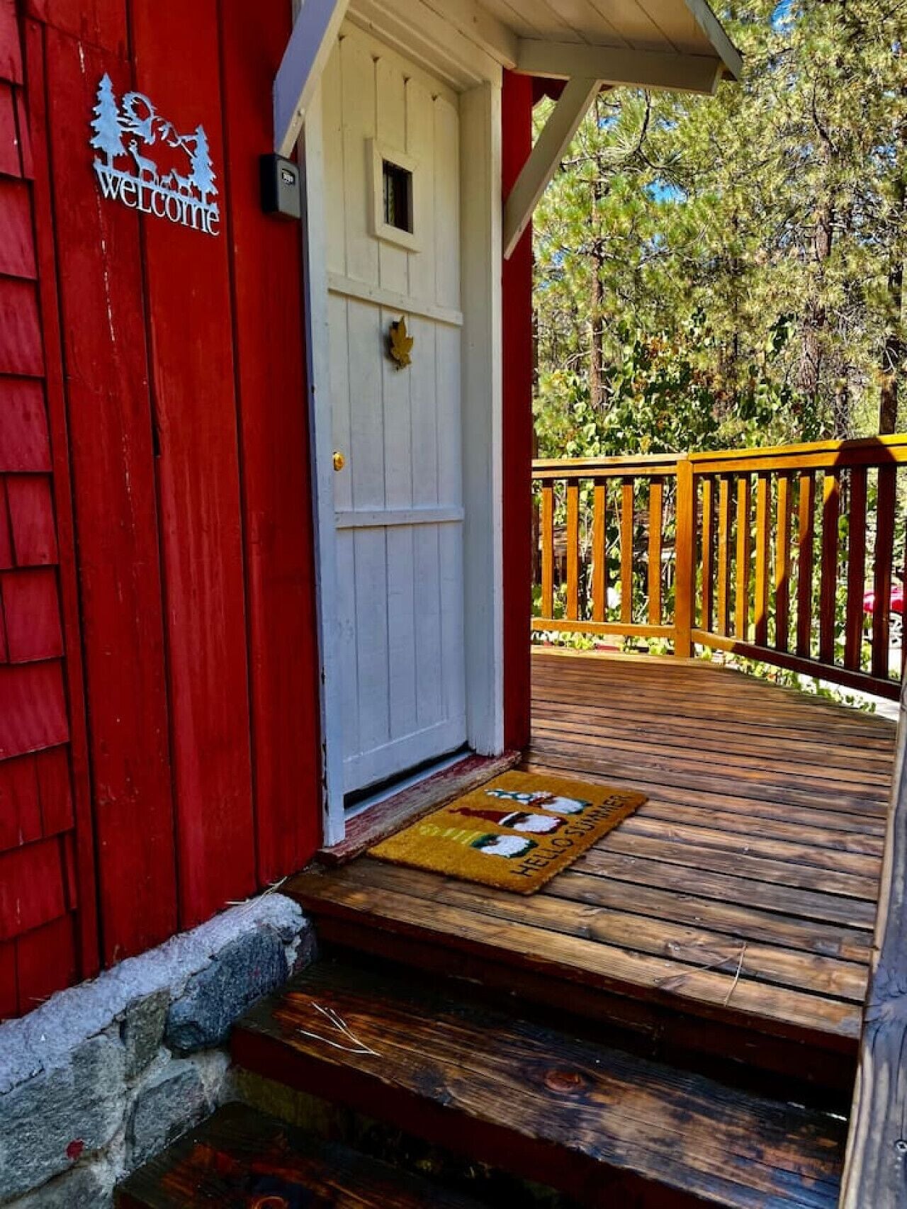 Un bâtiment rouge avec une porte blanche, une terrasse en bois et un paillasson jaune orné d’un motif représentant le Père Noël.