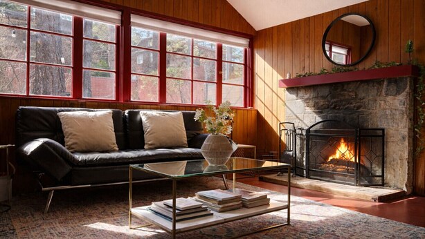 A cozy living room with a leather sofa, a glass coffee table with books, a fireplace, and large windows with red frames. - Peaceful Pines Stuga Near Ski Resort & Village (Wrightwood)