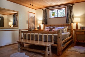A rustic bedroom with a wooden bed, a bench, a nightstand, a lamp, and a window with curtains.