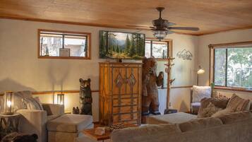 A cozy living room with a wooden ceiling, a ceiling fan, a large window, and a bear statue.