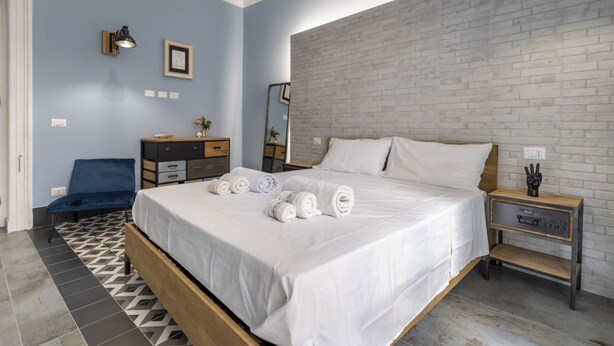 A bedroom with a bed, a nightstand, a dresser, a chair, and a wall with a brick pattern. - GuestHost - Cozy apartment equipped with all comforts and able to accommodate up to 4 people. The flat is located in a very central position: we are in fact a 10-minute walk from Piazza Maria Immacolata, just over 15 minutes from the Archaeological Museu (Taranto)