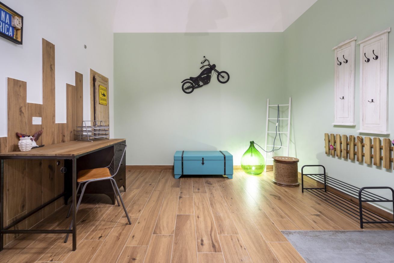 Et værelse med et træskrivebord, en blå opbevaringskasse, en motorcykelpynt på væggen, en grøn lampe og et stativ med kroge.