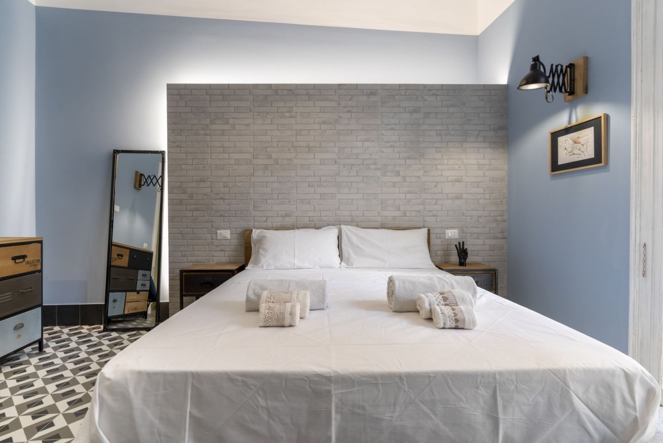 A bedroom with a brick-patterned headboard, a bed with white linens, a dresser, a nightstand, and a wall-mounted lamp.