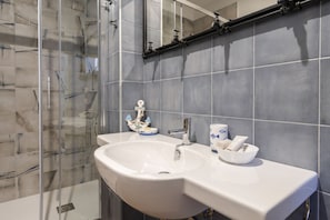A bathroom with a glass shower enclosure, a white sink with a mirror above it, and a tiled wall. - GuestHost - Cozy apartment equipped with all comforts and able to accommodate up to 4 people. The flat is located in a very central position: we are in fact a 10-minute walk from Piazza Maria Immacolata, just over 15 minutes from the Archaeological Museu (Taranto)