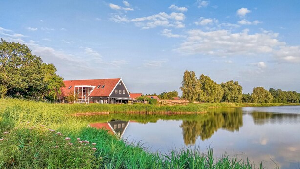 Ein Haus mit rotem Dach und großen Fenstern, neben einem stillen See mit Spiegelungen.