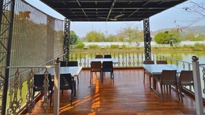 An outdoor dining area with tables and chairs, a covered roof, and a view of a river and trees. - Chang's Resort (Guwahati)