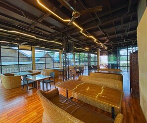 Un vaste espace intérieur doté de hauts plafonds, de poutres apparentes et d’un éclairage moderne. Des tables et des chaises sont disposées pour les repas ou les moments de convivialité.