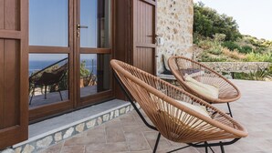 A stone building with large wooden doors and windows, wicker chairs on a tiled patio, and a view of the sea.