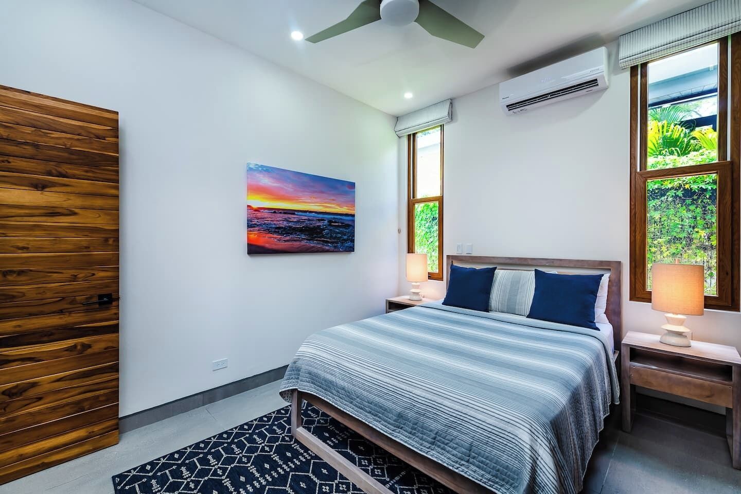 Un dormitorio con cama, mesitas de noche, ventilador de techo, un cuadro y ventanas con vistas a la vegetación.