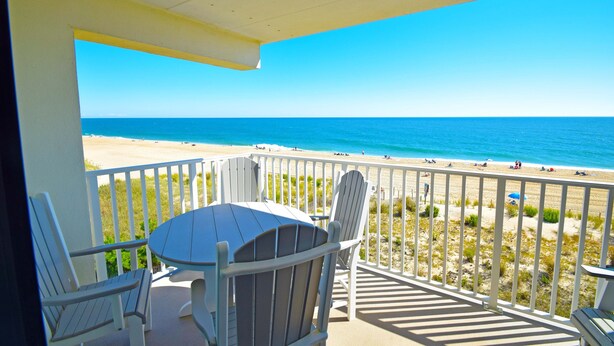 Un balcon avec vue sur la plage, des chaises blanches et une table.