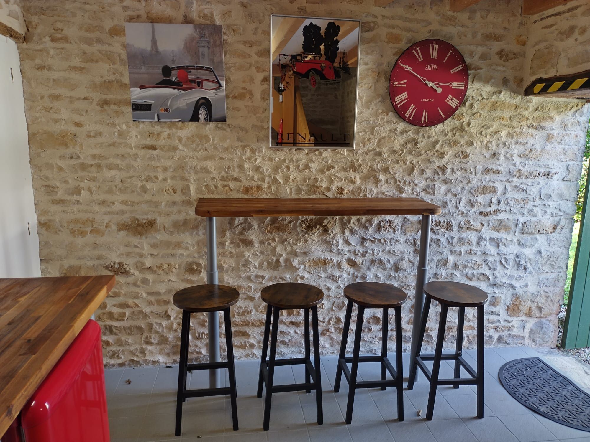 Un mur de pierres rustiques agrémenté d’un coin avec des tabourets de bar en bois, une horloge rouge et deux affiches.