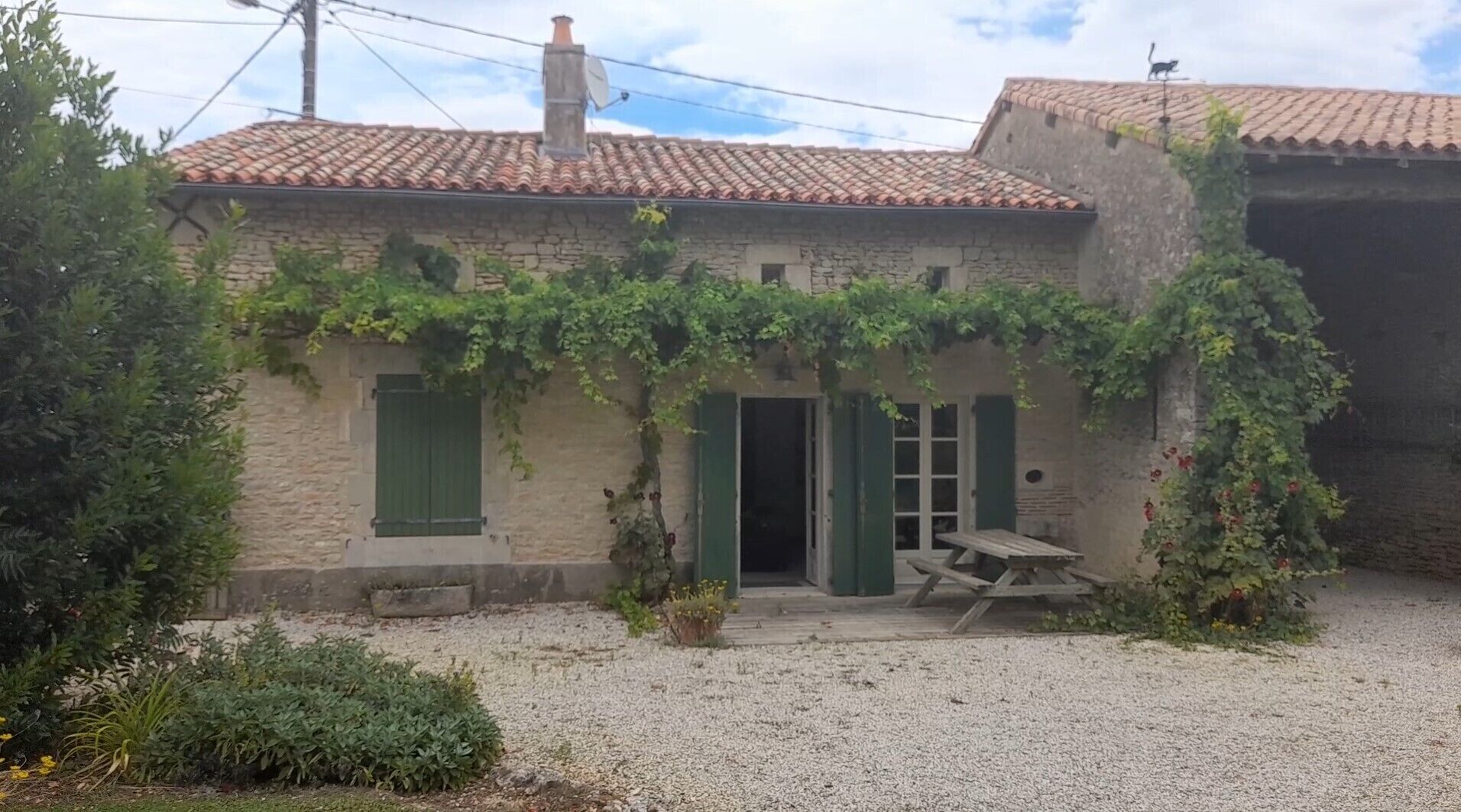 A rustic stone building with a tiled roof, green shutters, and a gravel driveway.