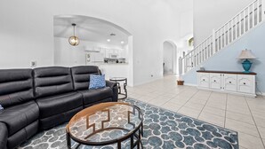 A modern living room with a black leather sofa, a glass coffee table, and a patterned rug. A kitchen is visible in the background with white cabinets and a blue accent wall. - Disney-Themed 5BR w/ Pool & Hot Tub • Sleeps 10 (Kissimmee)