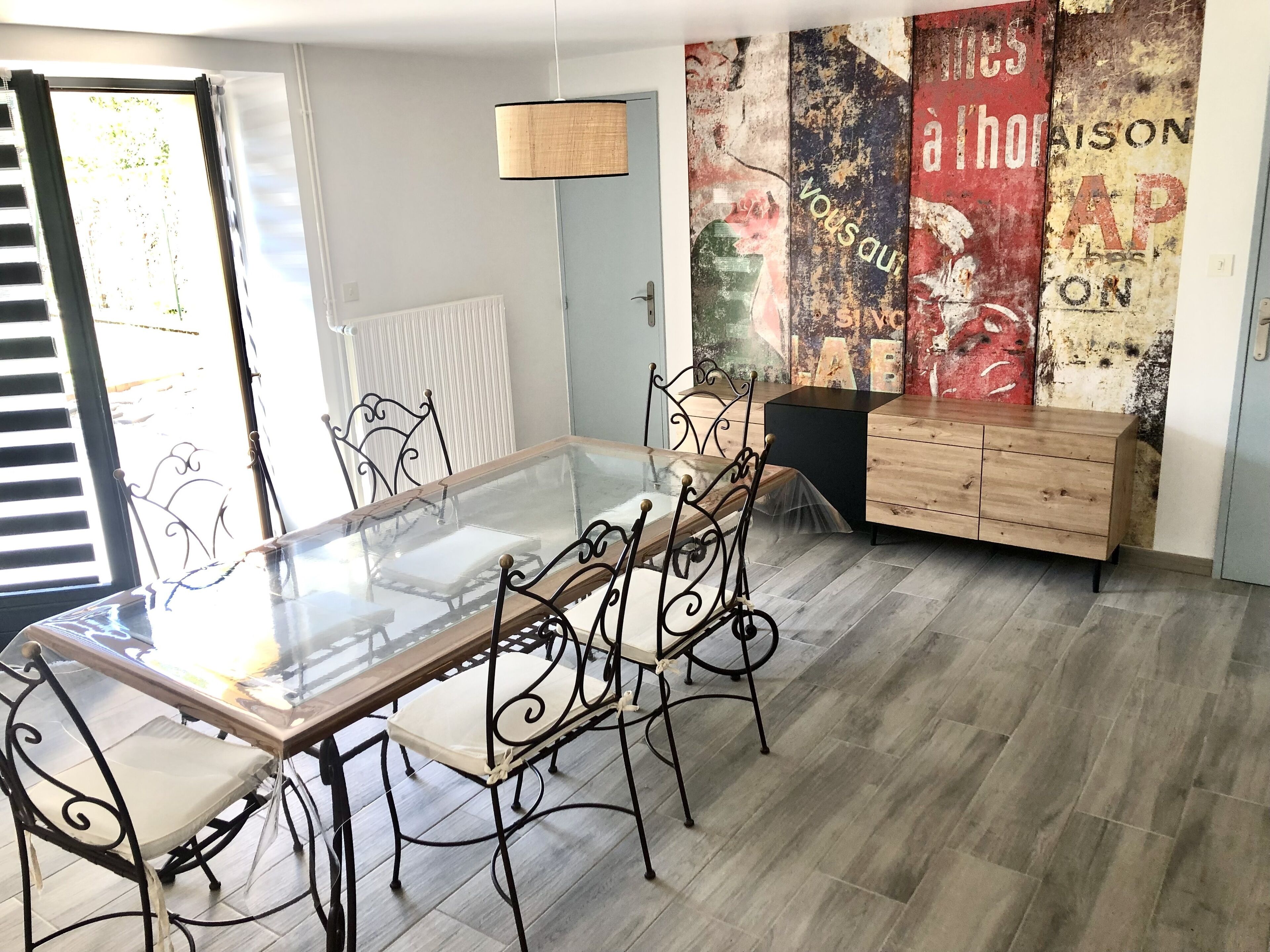 Un comedor con una mesa de vidrio, sillas de metal, un aparador de madera y una pared con carteles antiguos.