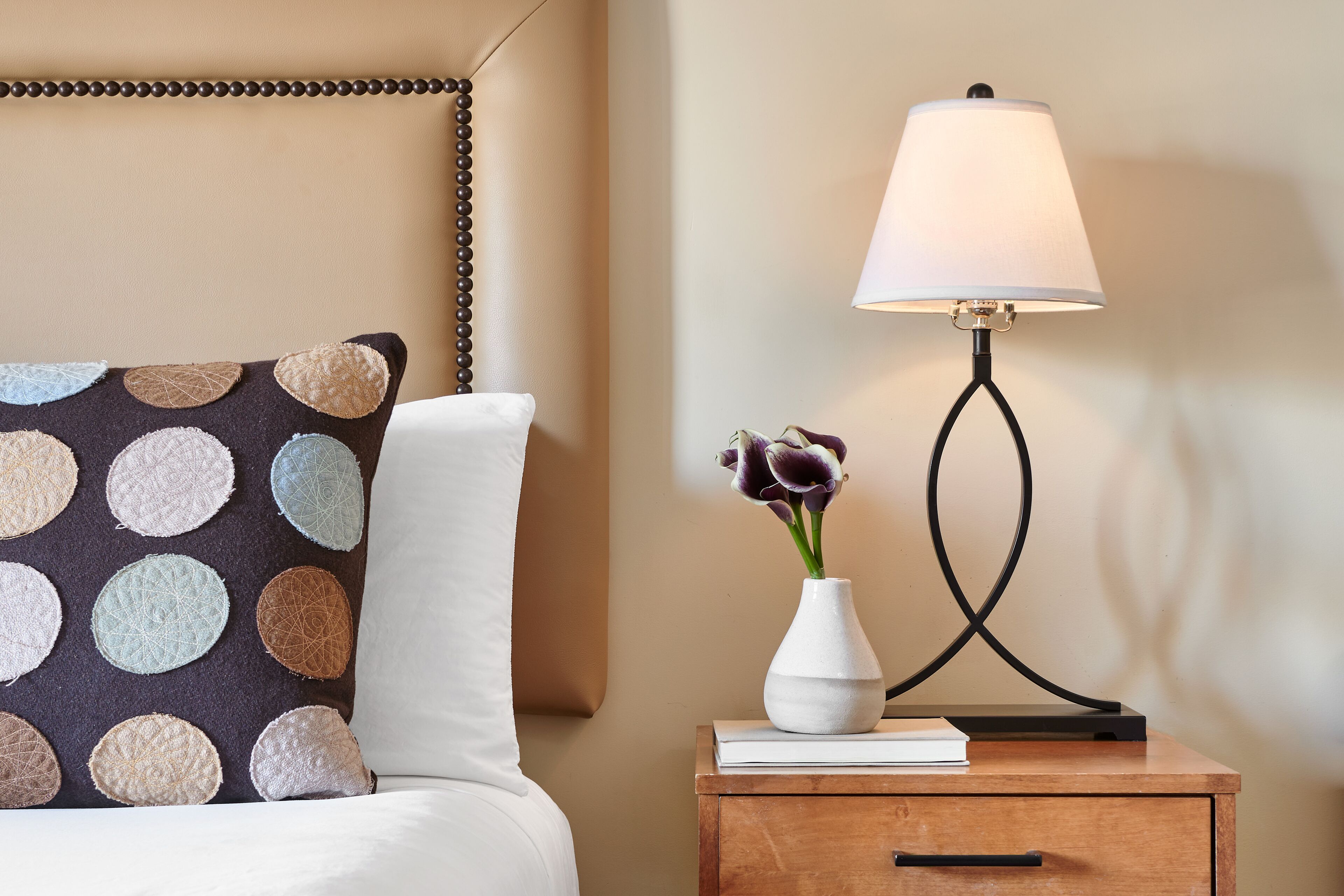 A neatly made bed with a patterned pillow, a bedside table with a lamp, and a vase with flowers.