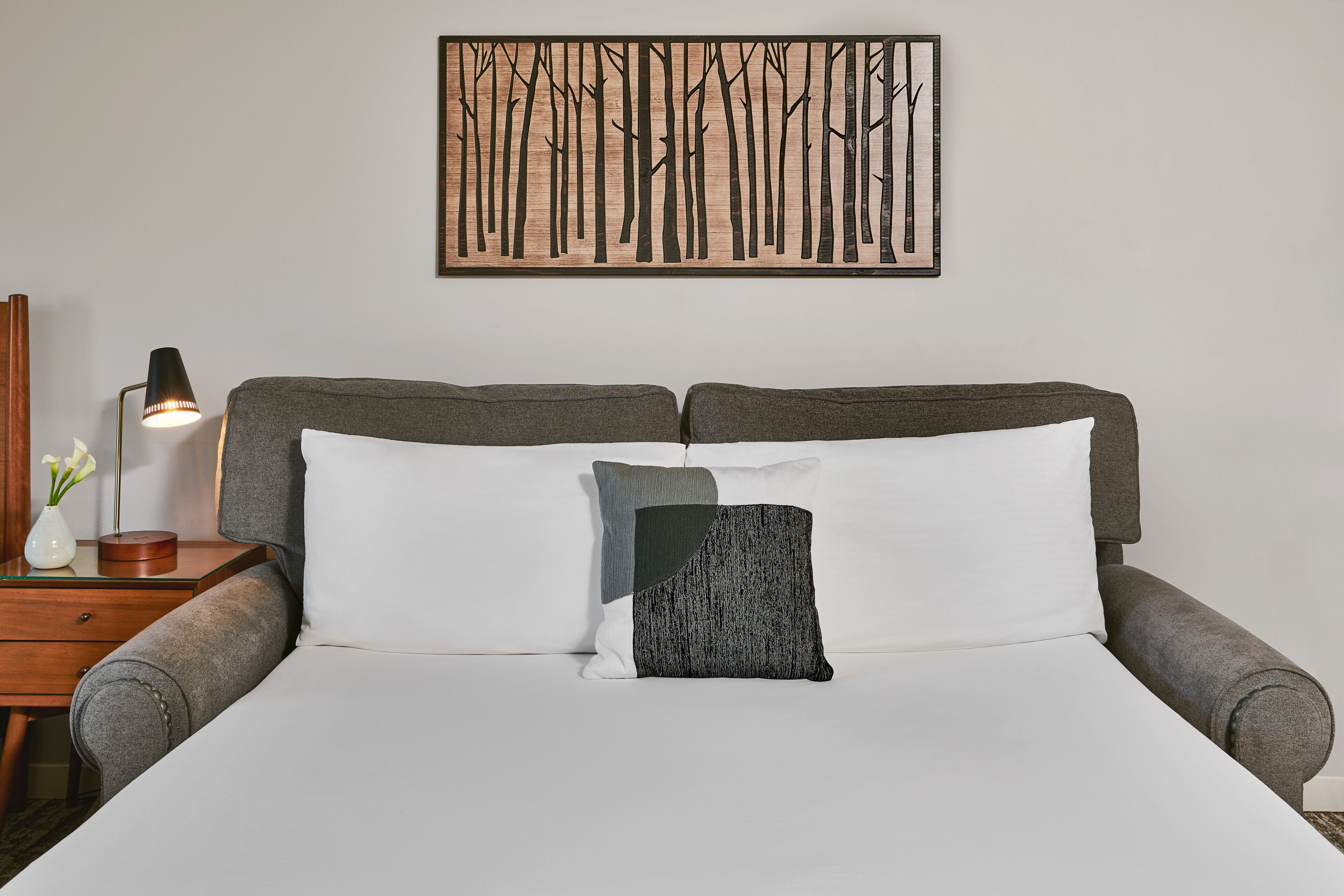 A neatly made bed with two pillows, a bedside table with a lamp, and a wall-mounted artwork of trees.