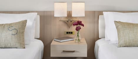 A hotel room with two beds, a nightstand with a lamp, a vase with flowers, and a book.