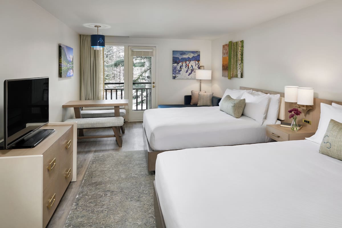 A hotel room with two beds, a TV, a desk, and a balcony with a view of trees.