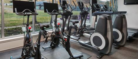 Une salle de sport équipée de vélos d’appartement et de tapis de course, dotée de grandes fenêtres et offrant une vue sur des arbres et une route.