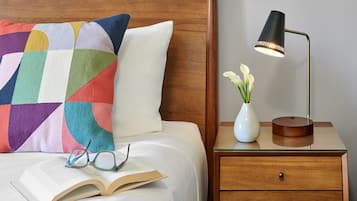A neatly made bed with a colorful pillow, a bedside table with a lamp and a vase of flowers, and an open book with glasses on it.