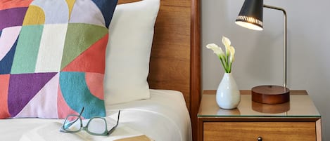 En pænt redt seng med en farverig pude, et sengebord med en lampe og en vase med blomster, samt en åben bog med briller på.