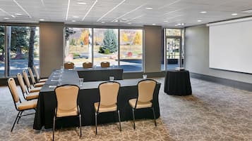 A conference room with a long table, chairs, a projector screen, and large windows with a view of trees and a building.
