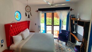 A bedroom with a red headboard, a bed with white linens, a dreamcatcher, a ceiling fan, a bookshelf, and a view of greenery outside.