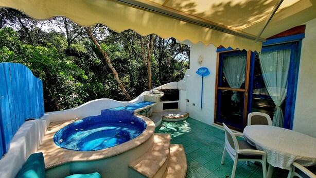 A pool area with a hot tub, a table, and chairs, surrounded by greenery.